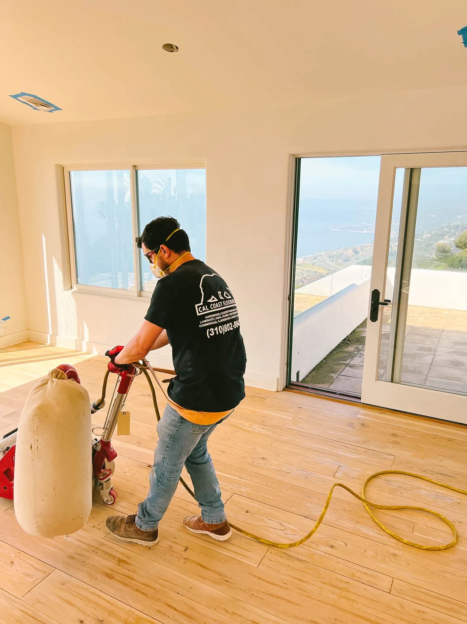 Cal Coast Flooring technician sanding a bare white oak floor with a drum sander, ocean view through patio doors in a Malibu home